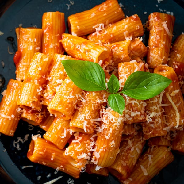 Rigatoni pasta with red sauce, grated cheese, and fresh basil on a dark plate.