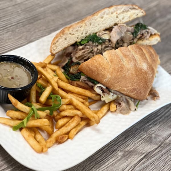 Italian beef sandwich with melted cheese and greens, served with fries and a side of au jus.